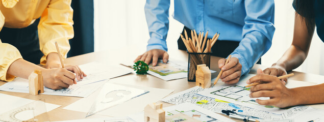 A portrait of professional architect team discussion about architectural project on meeting table with blueprint and wooden block scatter around at modern office. Closeup. Focus hand. Delineation.