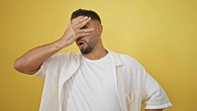 Startled young arab man with covering hand over face, peeking through fingers in shock - afraid expression on isolated yellow background