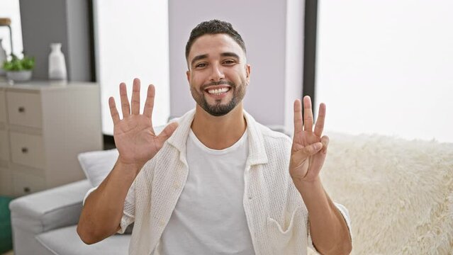 Cheerful young arabian man flaunting number eight with fingers, ecstatically pointing up on sofa indoors at home, embodies joy and positive vibes!