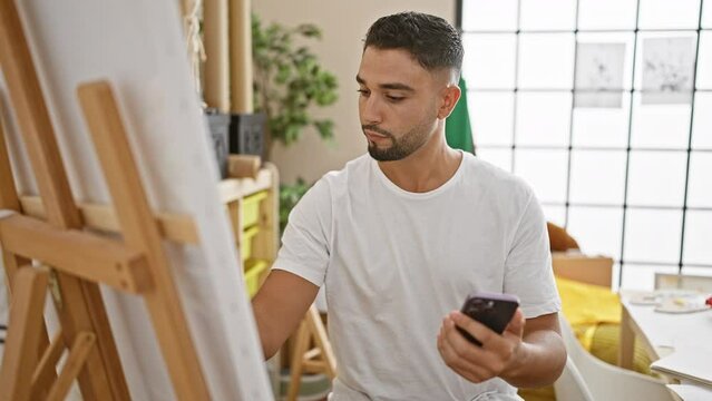 Focused man using smartphone while painting on a canvas in a bright art studio