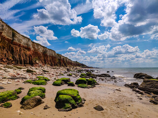 Scenic beach view of white and orange chalk and limestone cliffs at Old Hunstanton, Norfolk, England. Sand, sea, blue sky and green rocks covered in seaweed