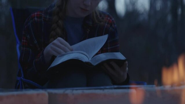 Young smiling woman sitting beside a camp fire and reading her Bible after sunset 