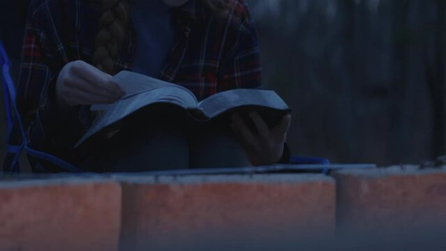 Young smiling woman sitting beside a camp fire and reading her Bible after sunset 