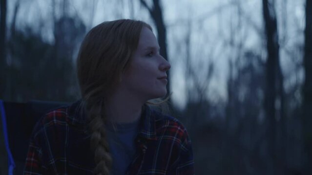 Young smiling woman sitting beside a camp fire and reading her Bible after sunset 
