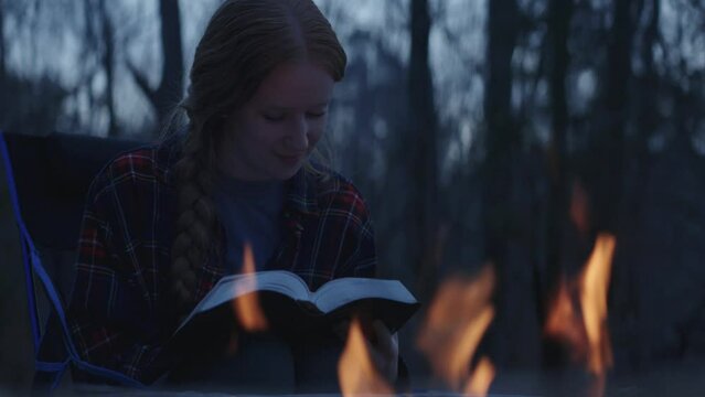Young smiling woman sitting beside a camp fire and reading her Bible after sunset 