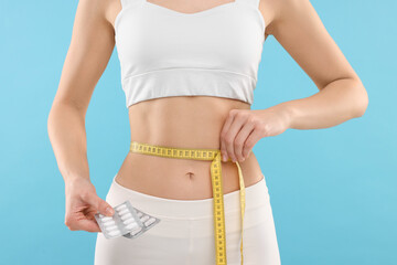 Slim woman holding pills and measuring waist with tape on light blue background, closeup. Weight loss