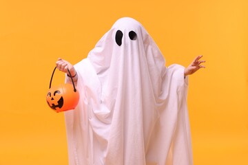 Child in white ghost costume holding pumpkin bucket on orange background. Halloween celebration