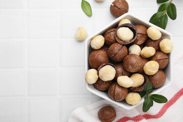 Tasty Macadamia nuts in bowl and green twigs on white tiled table, flat lay. Space for text