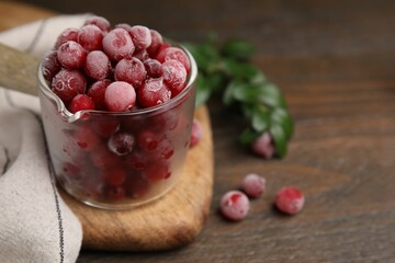 Frozen red cranberries in glass pot and green leaves on wooden table, closeup. Space for text
