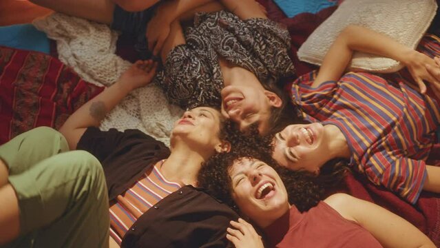 Company of young joyous girlfriends lying head to head on bed and laughing together when having fun at home party. View from above