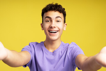 Portrait smiling boy, teenager wearing dental braces taking selfie isolated on yellow background....