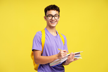 Smart smiling school boy wearing eyeglasses holding books taking notes isolated on yellow...