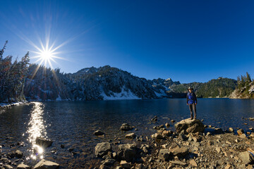 Adventurous athletic male hiker standing on a hill overlooking an alpine lake and tall mountains, on a beautiful sunny day in the Pacific Northwest.
