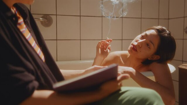 Young beautiful woman resting in a bathtub, smoking a cigarette and sharing with girlfriend sitting with book beside and reading aloud. Tilt-up shot