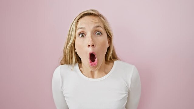 Young blonde woman, amazed and afraid, exhibits shock and excitement over isolated pink background