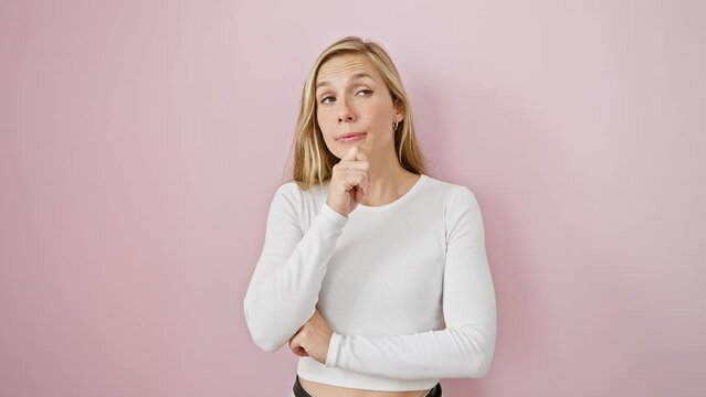 Thoughtful young blonde woman pondering a question, chin cradled in hand, a smile playing over her pensive face. confusion etched in her expression- an ambiguous puzzle on a pink isolated background.