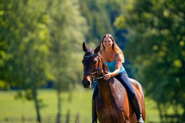 Obraz premium Young woman with brunette long hair rides bareback with her brown horse across a summer meadow, dressed in a blue tank top and riding pants with boots.
