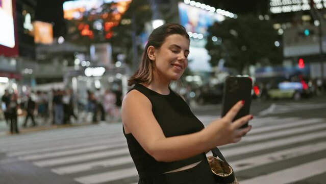 Smiling hispanic woman, casually snapping a beautiful selfie on tokyo's bright city streets at night, embodying the fun of urban technology tourism.