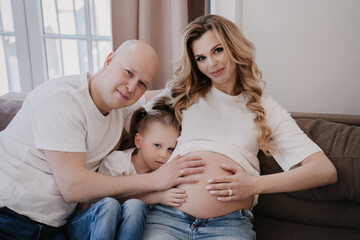 Intimate family portrait on a couch, with parents and child lovingly touching the mother's pregnant belly, anticipation in their eyes