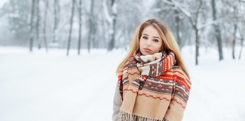 Beautiful fashion young blonde girl in a fashionable coat with a vintage scarf against the backdrop of a snowy park on a winter day
