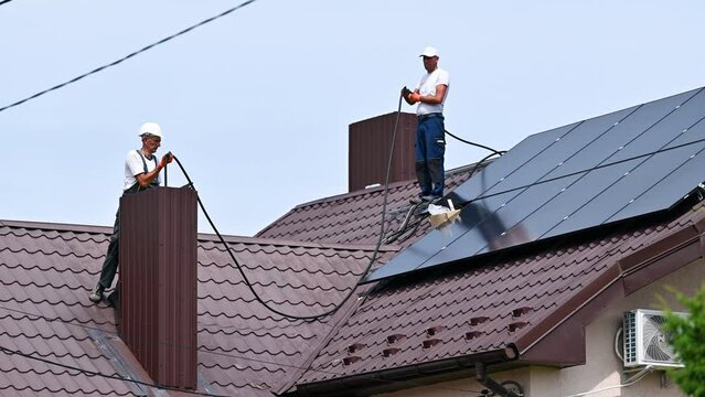 Workers installing solar panels system on rooftop of house. Electricians connecting cables from solar modules to inverter through the chimney for generating electricity through photovoltaic effect.