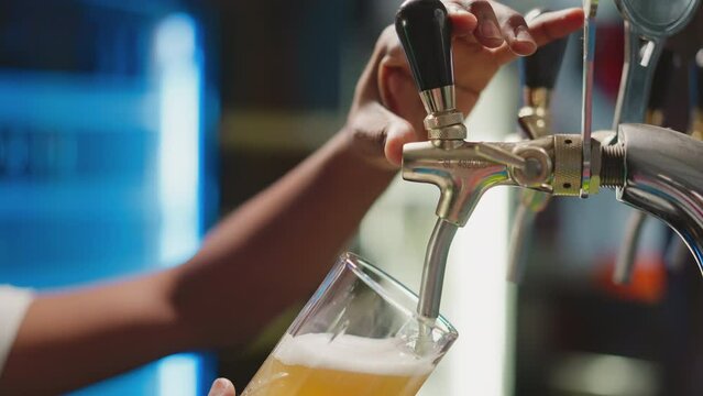 Barman fills mug with foamy beer from tap closeup. African American brewery worker pours fresh beverage into glass for guests. Man serves craft beer