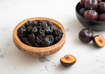 Two bowls with dried prunes and ripe raw plums on light background.