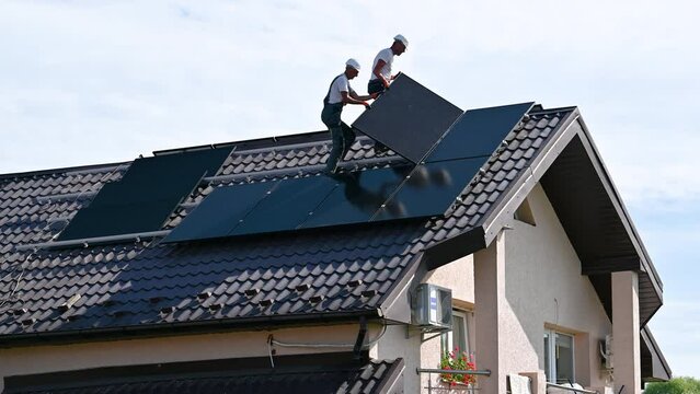 Workers building solar panel system on rooftop of house. Two men installers in helmets installing photovoltaic solar module outdoors. Alternative, green and renewable energy generation concept.