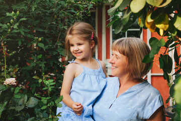 Loving middle-aged grandma holding smiling cute girl granddaughter while standing in garden