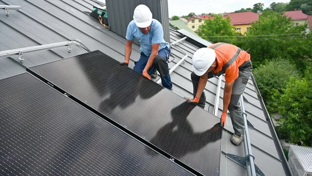 Workers building solar panel system on rooftop of house. Two men installers in helmets installing photovoltaic solar module outdoors. Alternative, green and renewable energy generation concept.