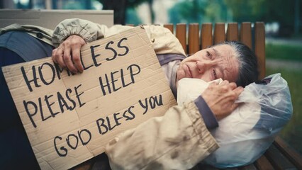 Desperate crying senior woman lying on bench, holding homeless cardboard sign