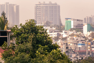 A hazy cityscape of modern concrete buildings beyond green trees.