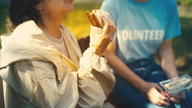 Pleased senior homeless woman eating sandwich, volunteer donating food, help