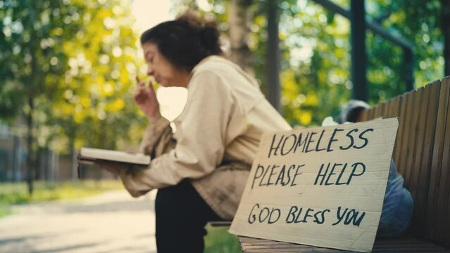 Homeless senior woman reading a book on bench, please help cardboard sign nearby