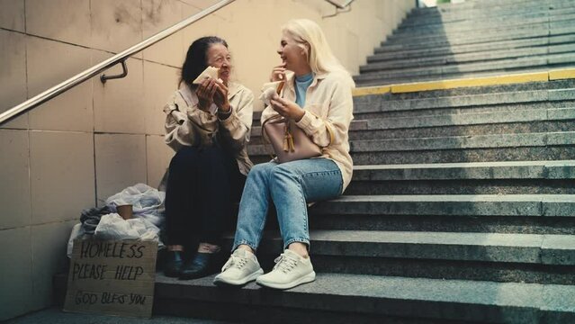 Thankful elderly homeless woman and woman passerby eating sandwiches, charity