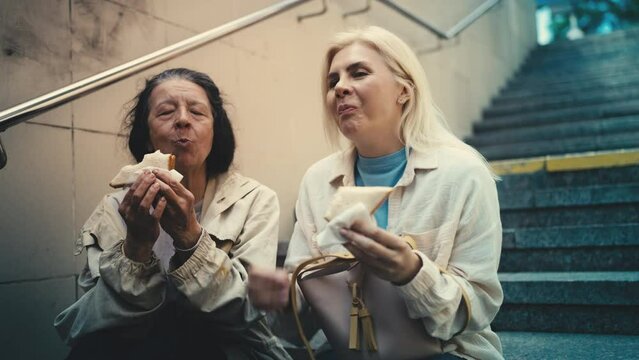 Pleased old female refugee and kind woman eating sandwiches together, charity