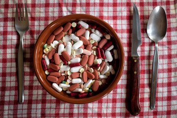 Vista superior de un plato repleto de pastillas junto a unos cubiertos, simbolizando el exceso en la ingesta de medicamentos - obrazy, fototapety, plakaty