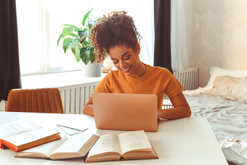 Cheerful young African American female student sitting at desk at home surrounded by textbooks, in front of laptop studying hard prepares for exams online,