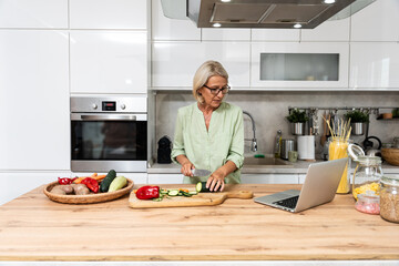 Senior woman a widow cooking vegetarian healthy food from vegetables at her home in domestic kitchen, using laptop computer for online recipes, preparing dinner for her date after long time