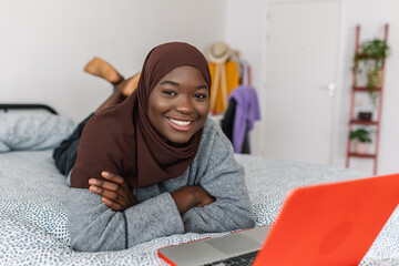 Joyful portrait of young teen african girl in muslim headscarf with laptop computer smiling at...