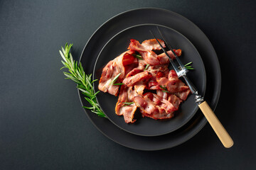  Roasted bacon slices with rosemary on a black plate.