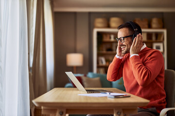 A cheerful male freelancer putting on his headphones to listen to a webinar related to his remote...