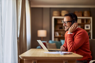 A cheerful adult male call center agent talking with a customer using headphones and a laptop.