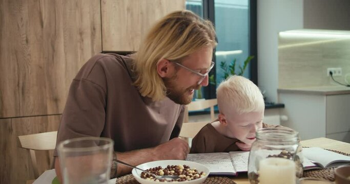 A little albino boy with white hair reads a book with his father, a blond man with glasses and a beard, in the kitchen after breakfast in the morning in a modern apartment