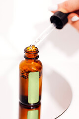 Extreme close up of womans hand and pipette. Transperent drop of essence, oil or gel drips from the pipette. Blurred brown glass serum bottle on the background. Concept of natural organic cosmeti