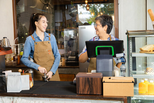 Young barista female hiring senior, elderly, 60s pensioner worker working in cafe bakery small business shop, two waitresses women standing behind coffee counter using technology cash register machine
