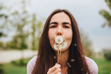 Portrait of young woman blowing dandelion outdoor in park on summer day