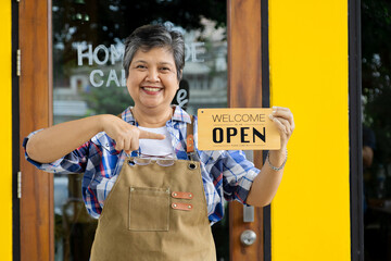 Asian 60s healthy senior barista worker female holding opening sign board pose in front of bakery...
