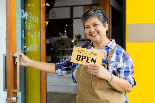 Asian 60s healthy senior barista worker female holding opening sign board pose in front of bakery shop retail small business owner start-up routine working, happy elderly retired pensioner lifestyle