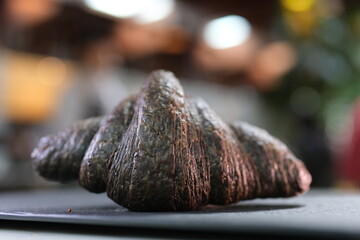 black croissant with cuttlefish ink on a white plate with a cup of cappuccino latte coffee against a green window background summer breakfast, France gourmet food delicious lunch snack